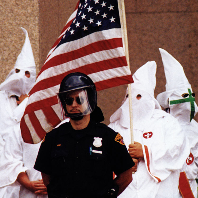 a mustachioed cop in riot gear wearing aviator sunglasses stands guard in front of several Ku Klux Klan members in full robes, one of whom carries a American flag