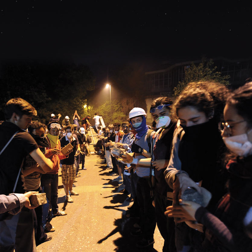 at night, lit by streetlights, young protestors in Turkey form a fire-brigade-line to transport cobblestones to the front lines of a confrontation with police